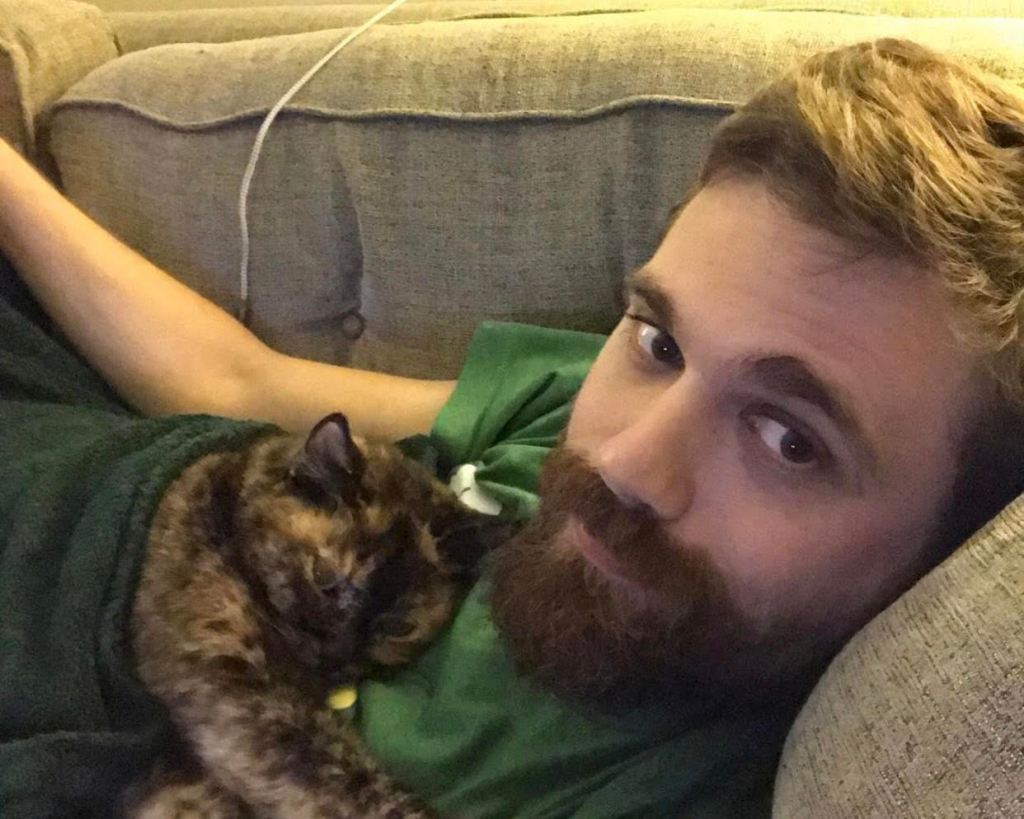Selfie photo, Person wearing green T-shirt, black/brown beard and moustache, lying on sofa, grey/brown coloured cat close to chest, arms stretched out across chest] 