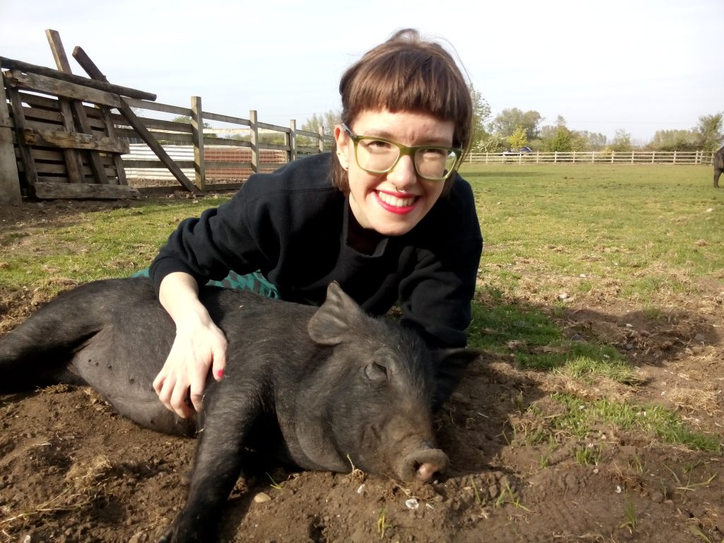 Person wearing glasses, red lipstick, in pasture, lying on ground, bent over brown/black pig, pigs has eyes closed and seems to be enjoying a belly rub 